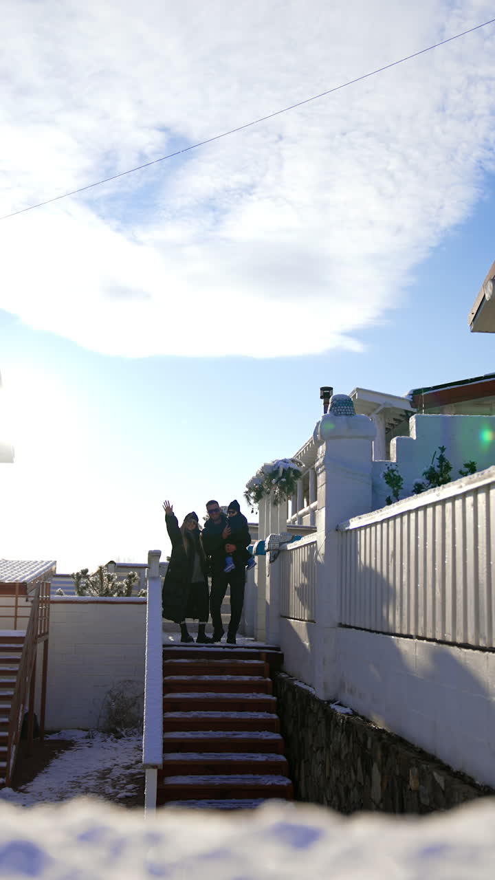 Young parents with little baby stands at the stairs outdoors. Family wearing warm clothes wave to camera. Vertical video.