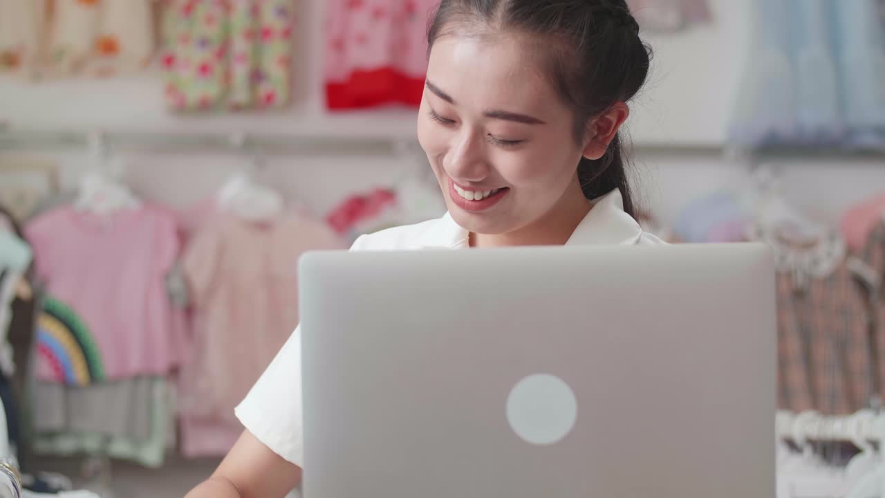 아시안 여성 어린이 의류 가게 소유자가 라인에 있는 재고를 확인하기 위해 노트북을 사용하는 것을 즐긴다.