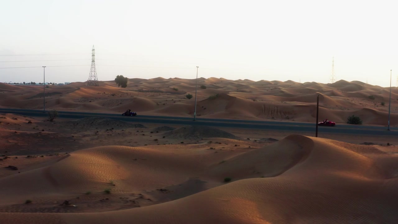 Vista laterale da drone che segue due veicoli in viaggio lungo una strada circondata da dune di sabbia illuminate dal sole nel deserto di Dubai, Emirati Arabi Uniti
