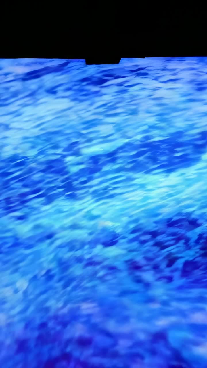 Video verticale: riproduzione su schermo illuminato che mostra acqua blu che scorre in una stanza buia con notch nero