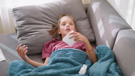 sick girl lying on couch covered with blue blanket, holding tissue with tissues scattered around, looking unwell, showing signs of cold or flu, resting and recovering from illness