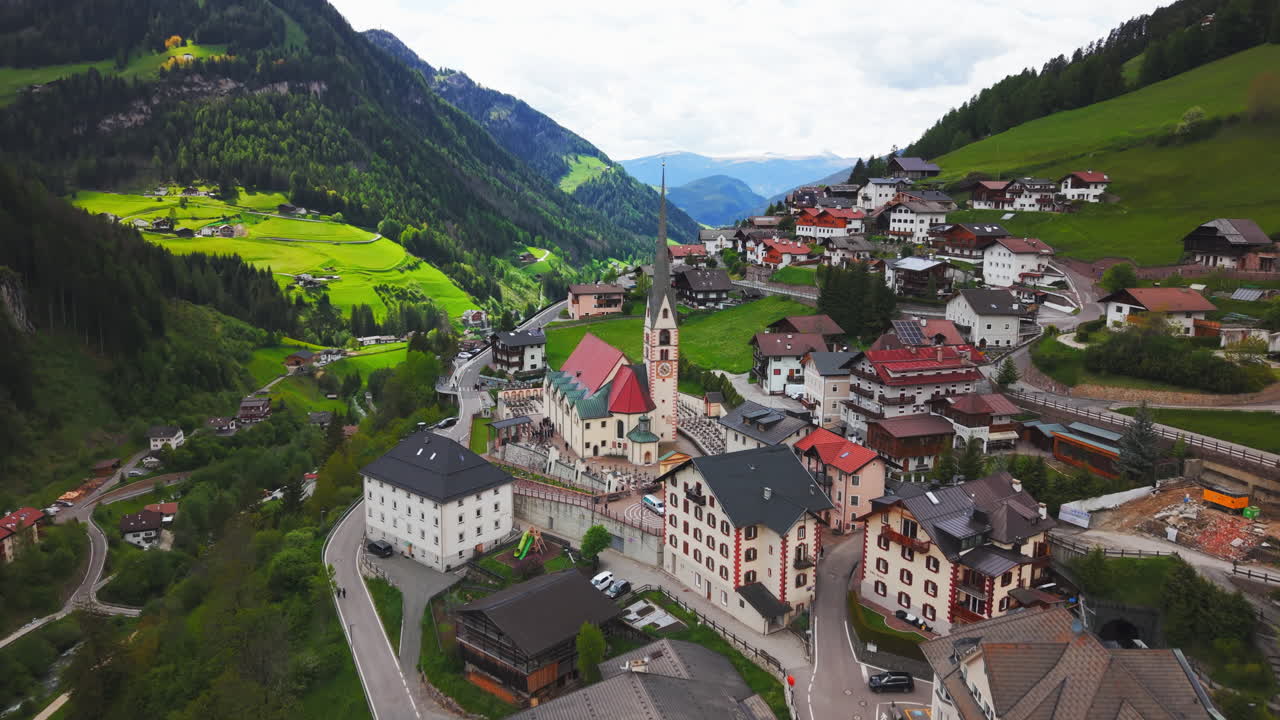 Drone footage of a slow rising aerial shot transitioning to a top-down view of a church with red roof and surrounding alpine village in the Dolomites, Italy
