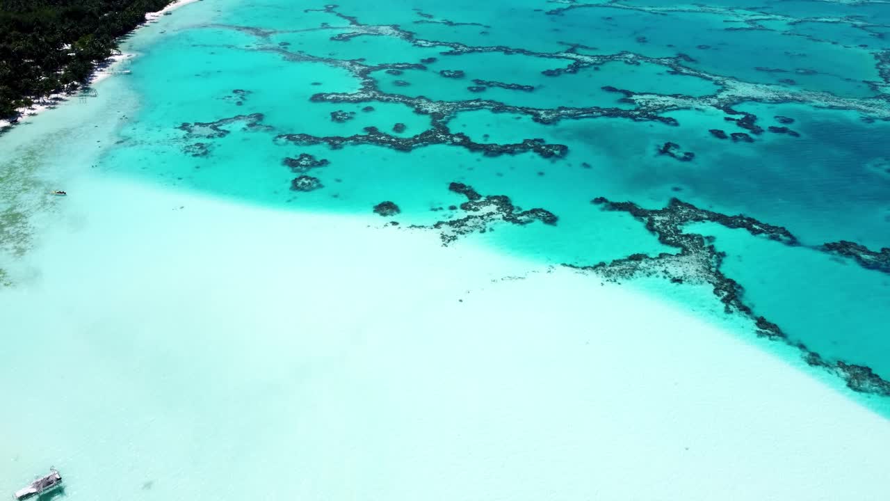 Drone view of a white sand beach, shallow water, coral reef and a green tropical pacific island with a lagoon on a sunny day in Maupiti, French Polynesia.