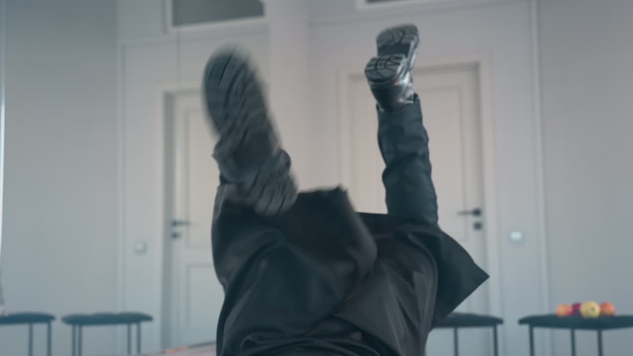 melodramatic male dancer in long black coat begins to fall toward ground mid-performance, arms raised expressively, dramatic movement framed by minimalist white studio background