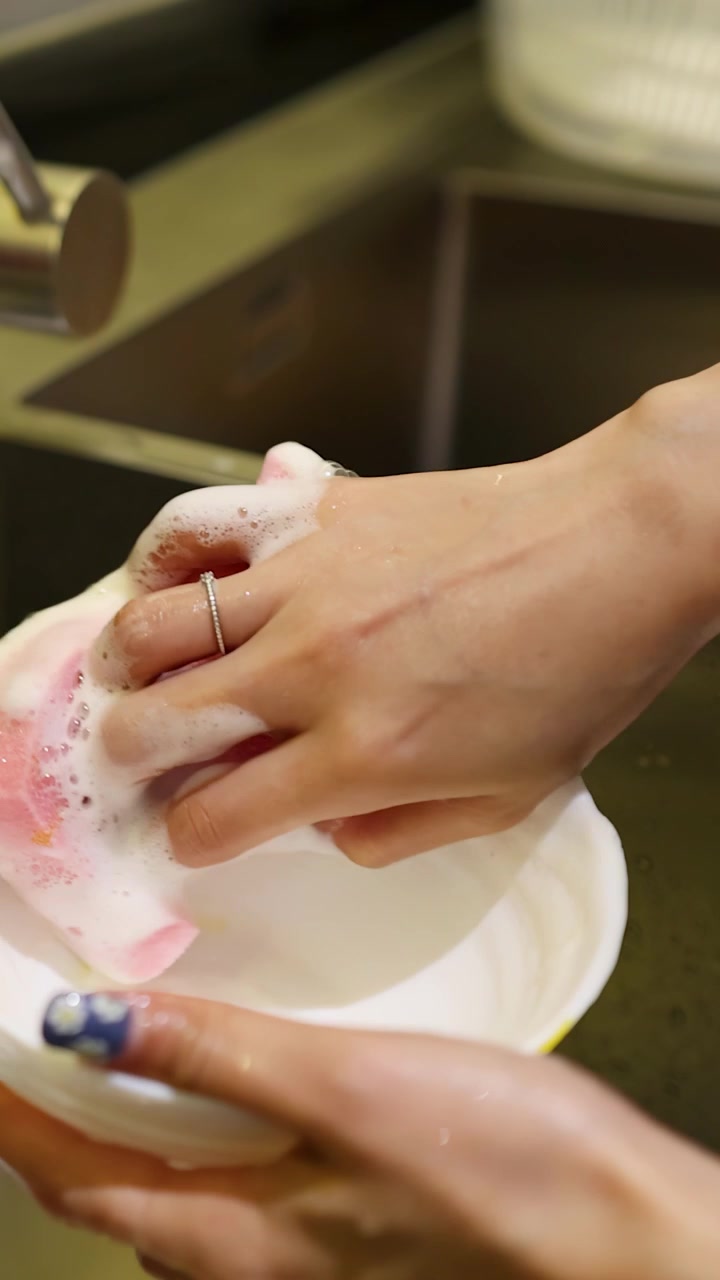 Primo piano di mani che strofinano una ciotola bianca con una spugna rosa e acqua saponata.