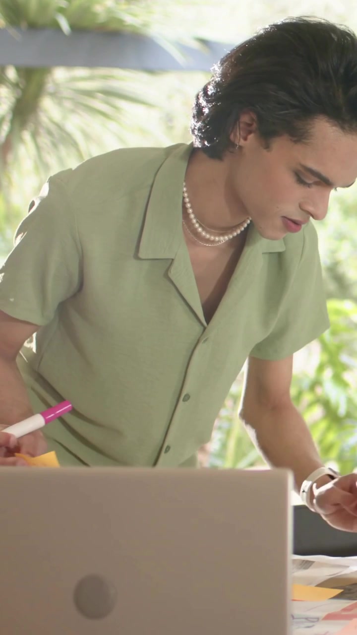 Video verticale: Un uomo con una camicia verde segna documenti a una scrivania vicino a una finestra con un laptop, usando un evidenziatore rosa.