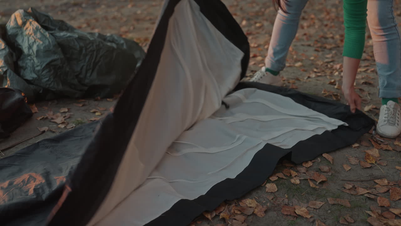 Fäller ut tältduk på löv, campare förbereder skydd i skymningen med fokuserade händer i jeans och sneakers, slätar ut innertältet, kontrollerar dragkedja och placering av tältstänger, fäster kanter med tältpinnar och testar.
