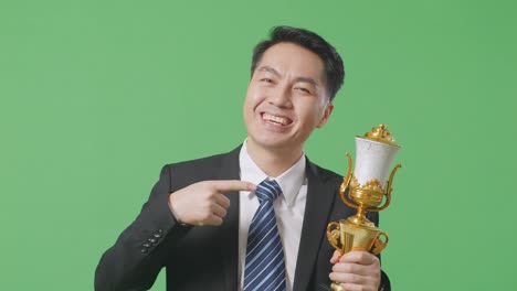 close up of asian business man in a suit and tie with a gold medal pointing to a gold trophy in his hand being proud winning as the first winner on green screen background in the studio