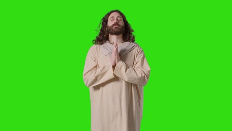 studio shot of man wearing robes with long hair and beard representing figure of jesus christ praying on green screen