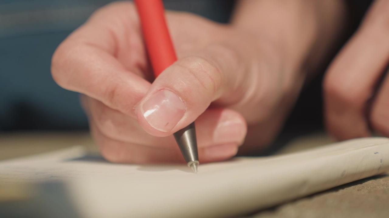 Primo piano di una mano che tiene una penna rossa e scrive su carta, con illuminazione soffusa e dettagli focalizzati. La mano si muove leggermente mentre scrive, mantenendo una stretta vicinanza al foglio bianco per tutto il tempo.