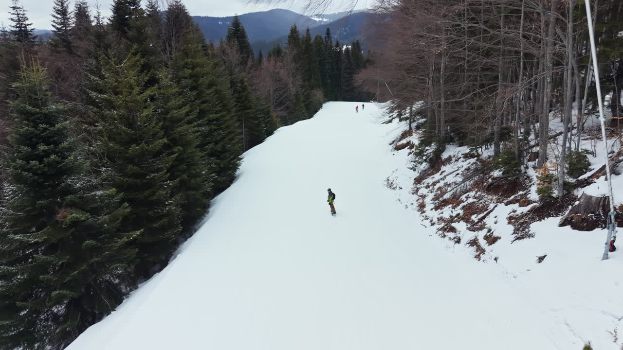 Uno snowboarder scende un pendio innevato in una zona boschiva durante l'inverno in montagna a mezzogiorno, vicino a piste da sci e altre persone.