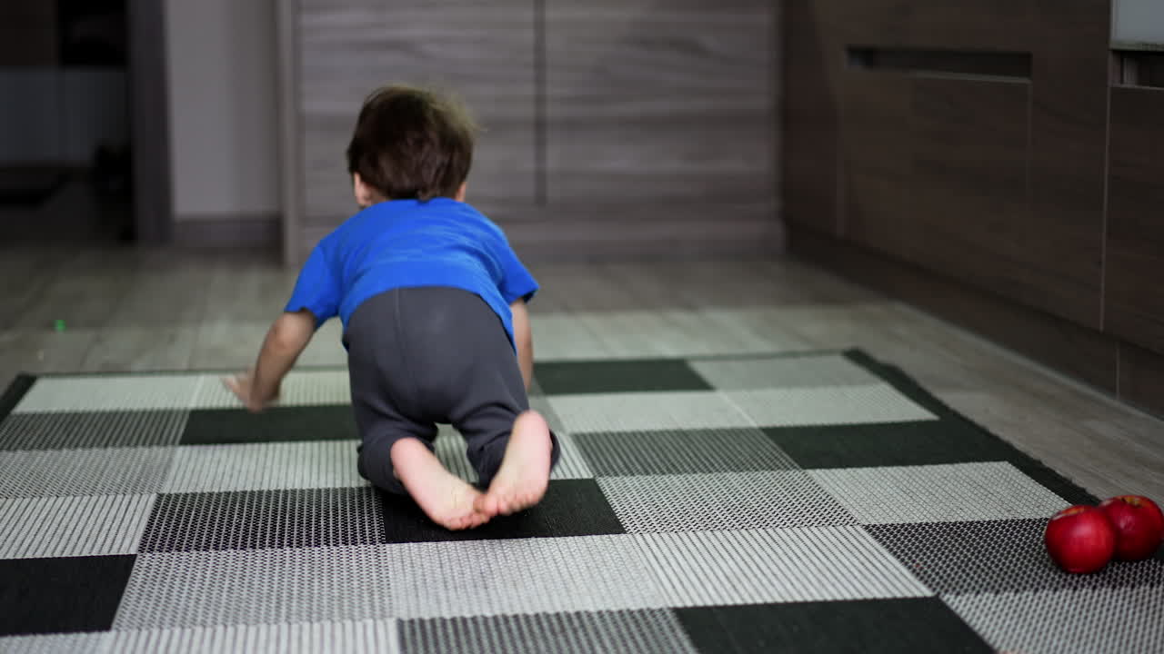 Little barefoot toddler sits on the carpet his back to camera. Baby boy crawls by the floor to pick an apple.
