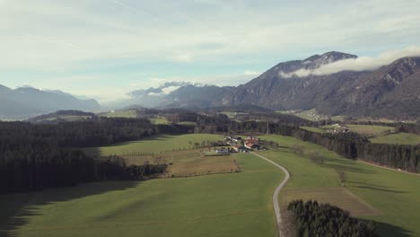 Luftdrohne-Der-Wunderschönen-Alpinen-Bergkette-In-Der-Ferne-Mit-Malerischen-Grasfeldern,-Bäumen-Und-Einer-Stadt
