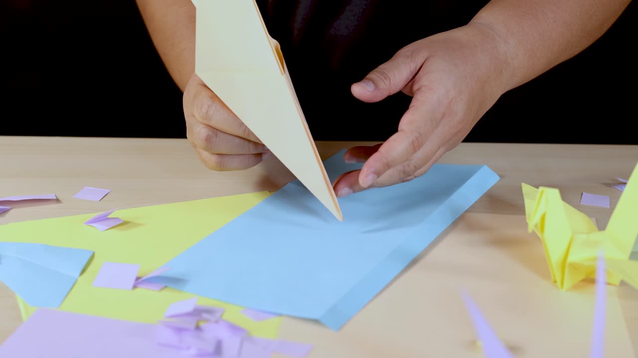 Person folds paper plane origami on desk with colorful paper, overhead lighting, steady camera