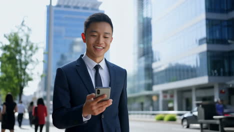 young asian businessman smiling while using smartphone outdoors