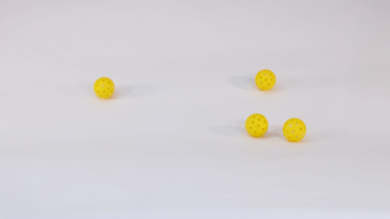 Three yellow plastic pickleball balls fall, bounce, and roll across a clean white studio surface