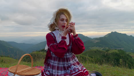 female blonde model in traditional old fashioned style clothing doing make up with lipstick and portable mirror in stunning outdoor mountains natural location