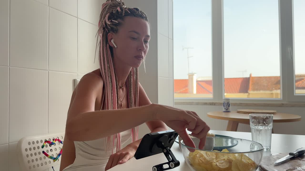 Giovane donna che mangia patatine e guarda il telefono in una cucina luminosa