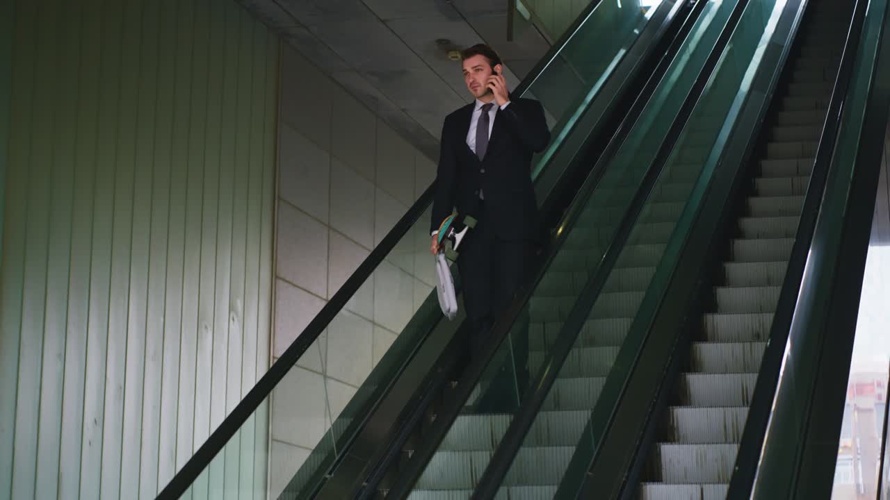 Plano de rastreamento de um profissional sorridente do sexo masculino, de terno e gravata, segurando um skate e usando o celular em uma escada rolante em um terminal corporativo moderno