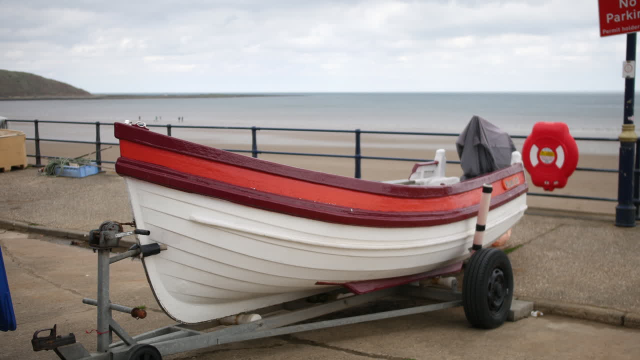 A boat with a white hull and red trim is resting on a trailer beside the beach promenade in Filey. The scene includes a life ring, railing, and ocean view under cloudy skies