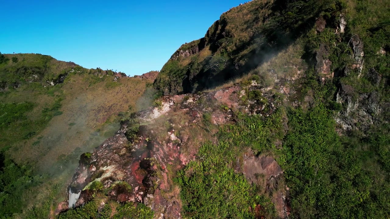 Vapore vulcanico che si innalza dal pendio del cratere con fumo leggero che si diffonde su un terreno accidentato, paesaggio vulcanico attivo con fumarole e vegetazione lussureggiante, riprese aeree con drone.