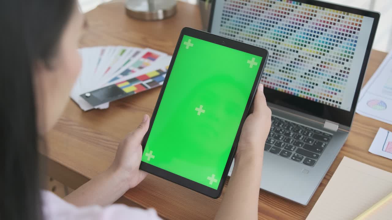 Creative designer Asian woman using digital tablet pad device blank green screen mock up, using computer laptop with color chart samples interior design, working in modern office workspace desk.