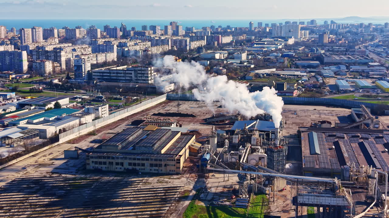 Fabrik i et industriområde sender røg op, mens bygninger og byens skyline er synlige i baggrunden i dagslys.