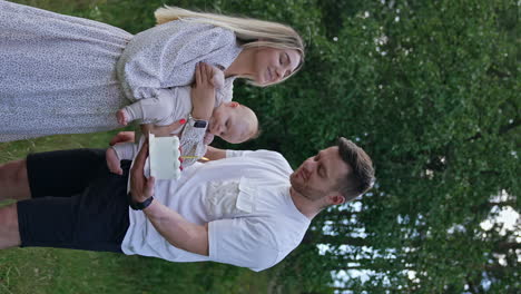 bearded man holding a cake in front of the woman with child in hands. parents make a wish and blow the candle on the cake. nature backdrop. vertical screen.