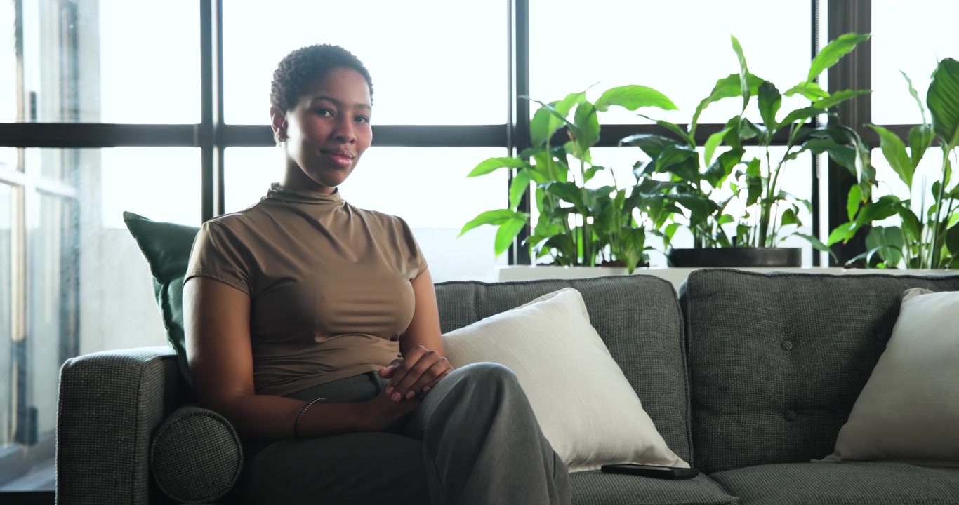 African woman scrolling smartphone and reading messages placing it on gray sofa in living room
