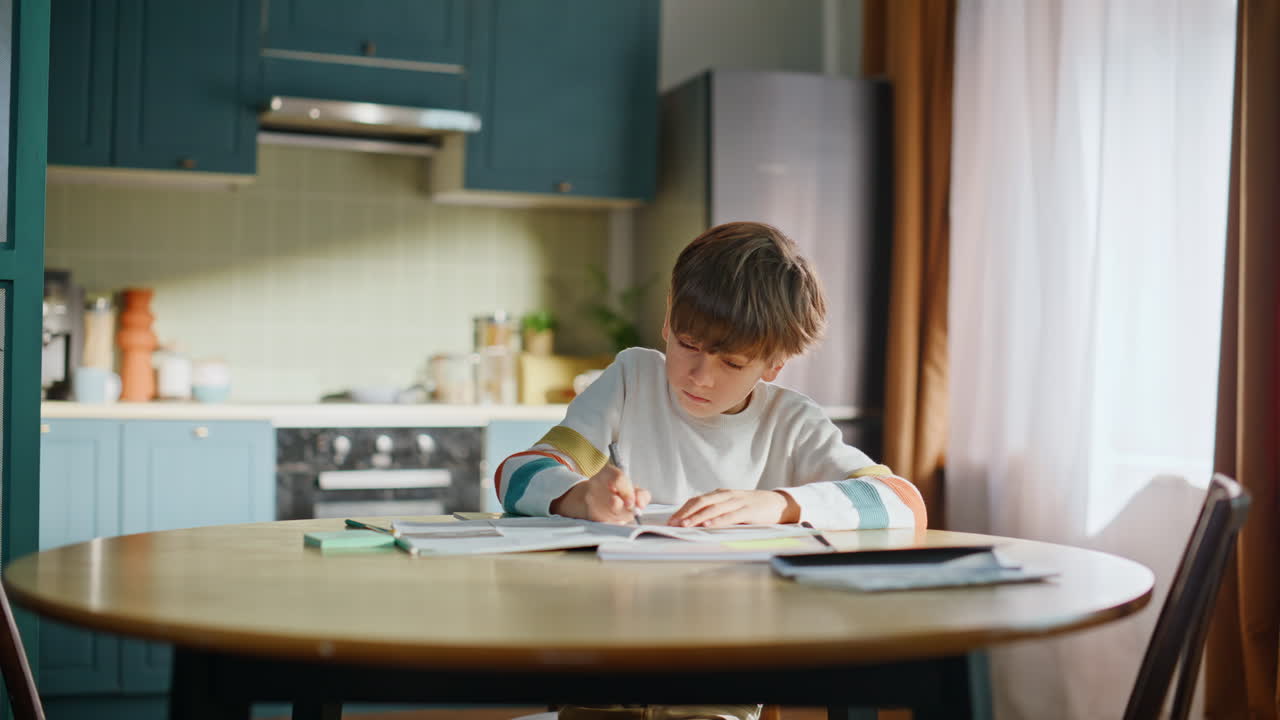 Smart schoolboy writing notebook making homework sitting kitchen table closeup
