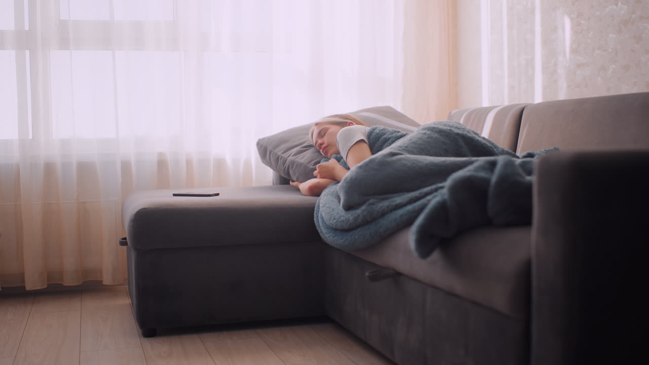 Independent lady sleeping on couch hugging blanket warmly with phone placed nearby peaceful expression on face as soft daylight streams through curtains creating calm cozy atmosphere of rest