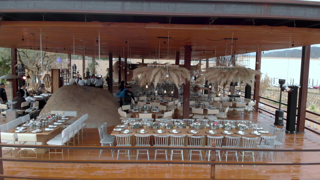 Drone-opname van een strandrestaurant in Mexico met lange eettafels, houten architectuur, hangende decoraties en grote open ramen met uitzicht op het kustlandschap