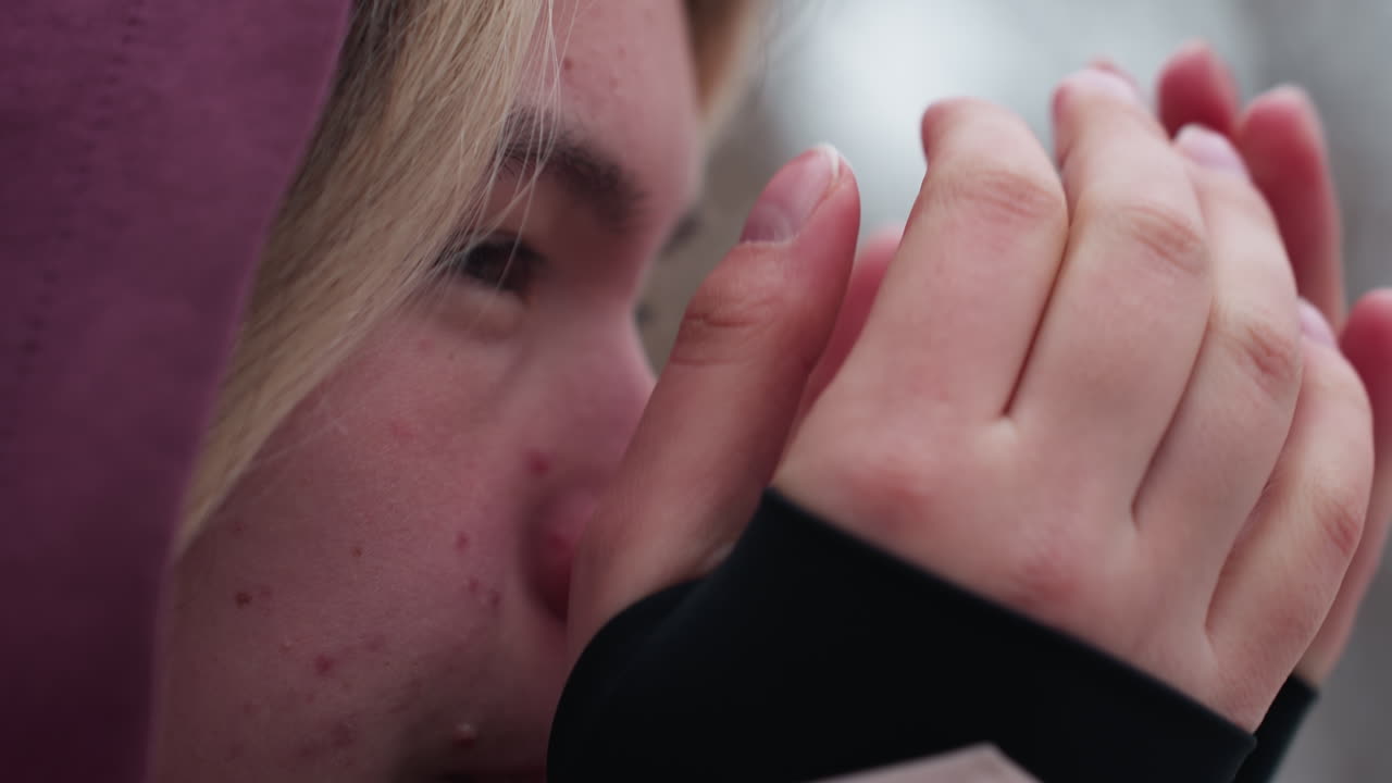 vista cercana del rostro de una mujer que siente frío en invierno, frotándose las manos para calentarse, nariz roja y piel agrietada muestran condiciones climáticas duras, fondo borroso de árboles desnudos