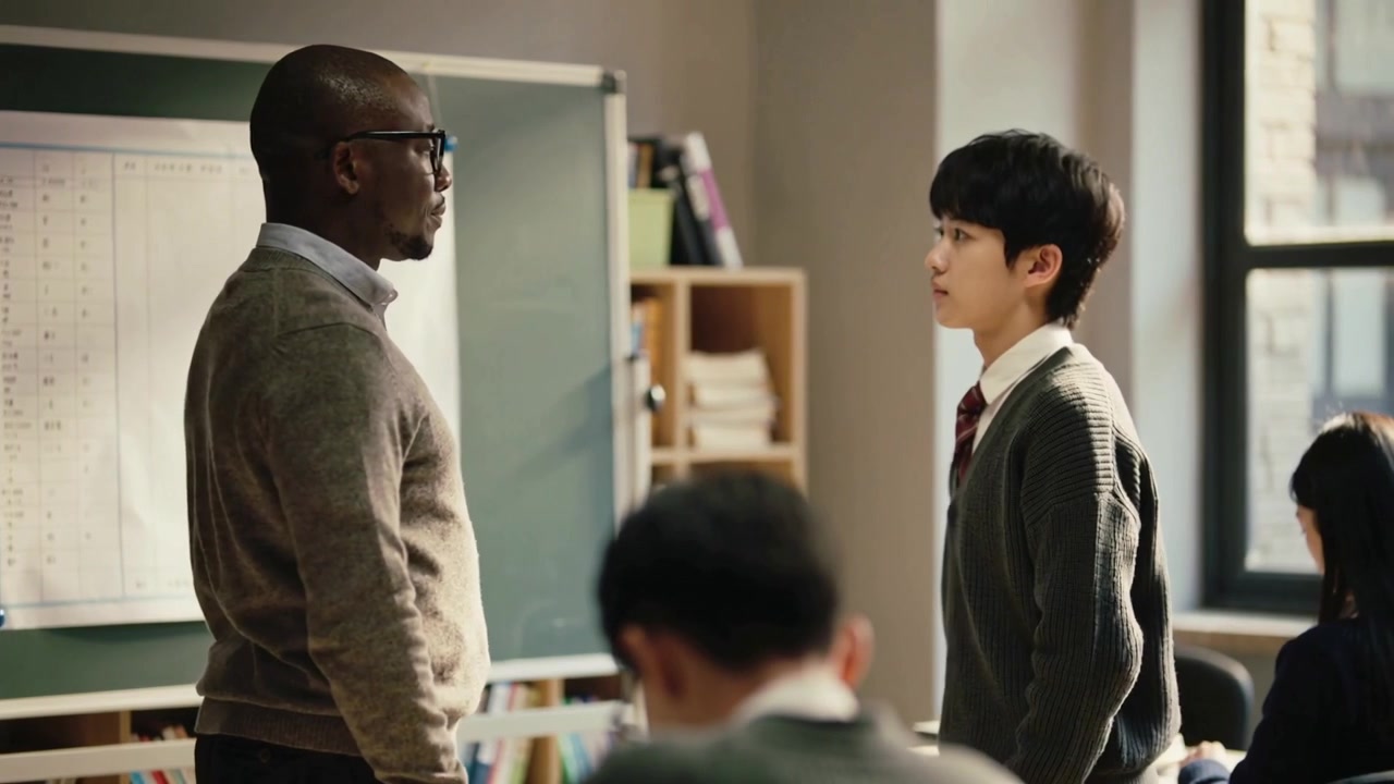 Rising student in uniform stepping toward teacher in sweater at classroom front by green board