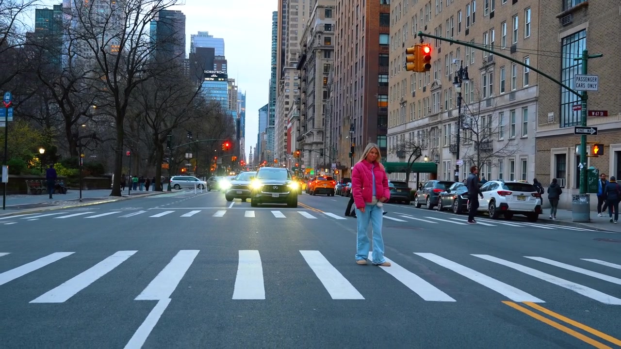 New York, USA, 24 March 2026: Woman in pink jacket crossing busy New York street. Pedestrian in pink clothing crossing an avenue with cars and traffic lights in Manhattan at twilight.