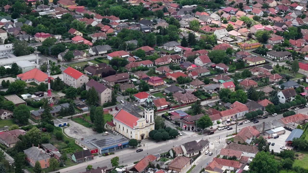 Et vidstrakt luftperspektiv viser en tradisjonell ungarsk by med røde tegltak, en sentral kirke og frodige grønne hager.