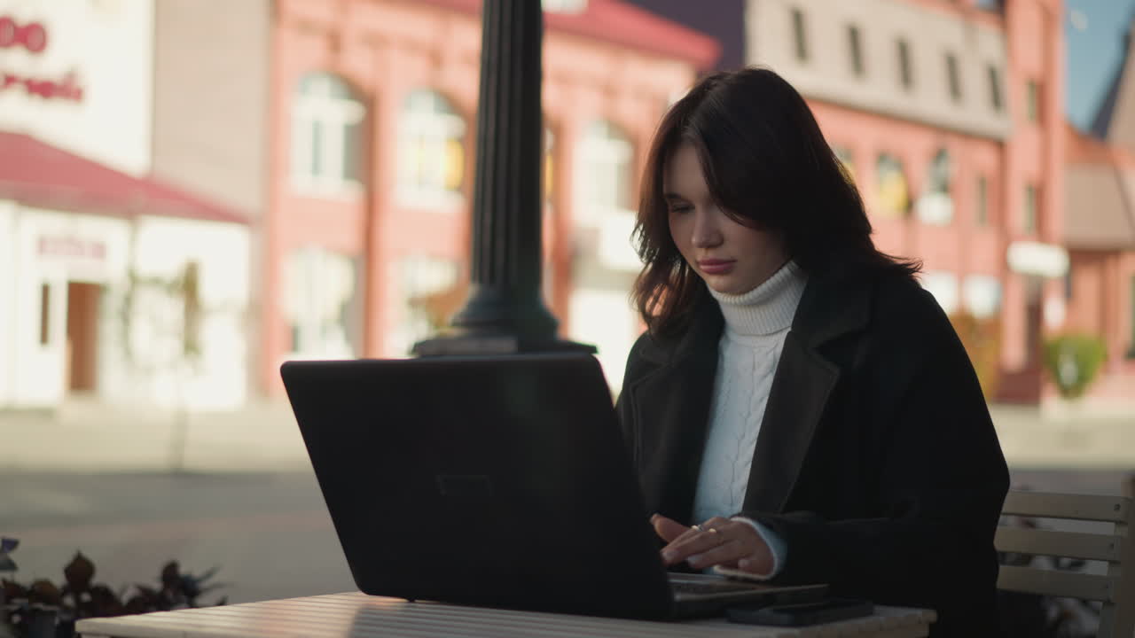 Young woman working on laptop outdoors in urban setting, wearing stylish black coat and white turtleneck, decorative black pillar nearby, blurred red brick building in background with shadows