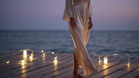 woman wearing flowing white dress standing on wooden pier beside sea during sunset, gripping skirt edges with soft string lights illuminating surrounding landscape