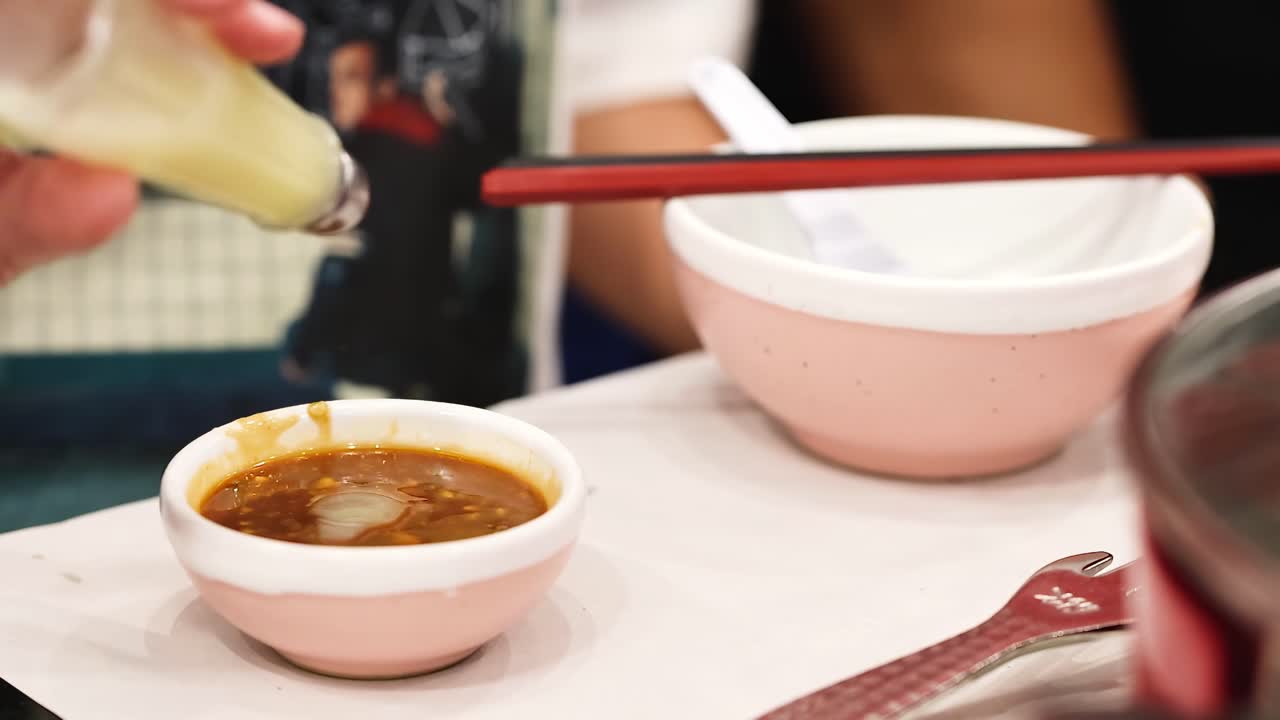 A hand pours sauce from a bottle into a small bowl beside chopsticks on a table.