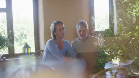 Smiling-senior-caucasian-couple-using-laptop-and-holding-documents-at-table