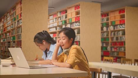 asian woman student with headphones thinking and looking around then raising her index finger while sitting with classmate writing into the notebook on table with a laptop in the library