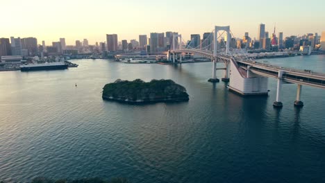 Drone-Aéreo-Volando-Bajo-Sobre-El-Agua-Hacia-El-Puente-Colgante-Del-Arco-Iris-En-La-Ciudad-De-Odaiba-Tokyo-Japón-Durante-La-Puesta-De-Sol