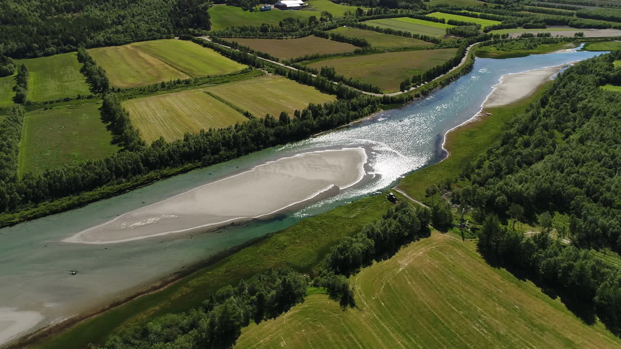 un dron volando en círculos sobre un río tranquilo con una isla de arena en medio de vibrantes campos verdes y áreas boscosas.