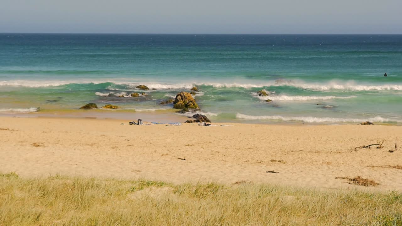 Slow motion landscape of ocean waves crashing on rocky boulders and seaside sandbar with beach towels and items laid on sand at beach in Yagon Seal Rocks in NSW Australia nature travel and outdoors