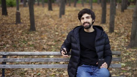 brunette man sits on bench in the autumn park, works on the phone