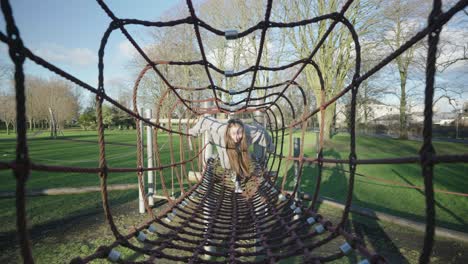 a young girl playing at the local park on the jungle gym made from rope surrounded by trees