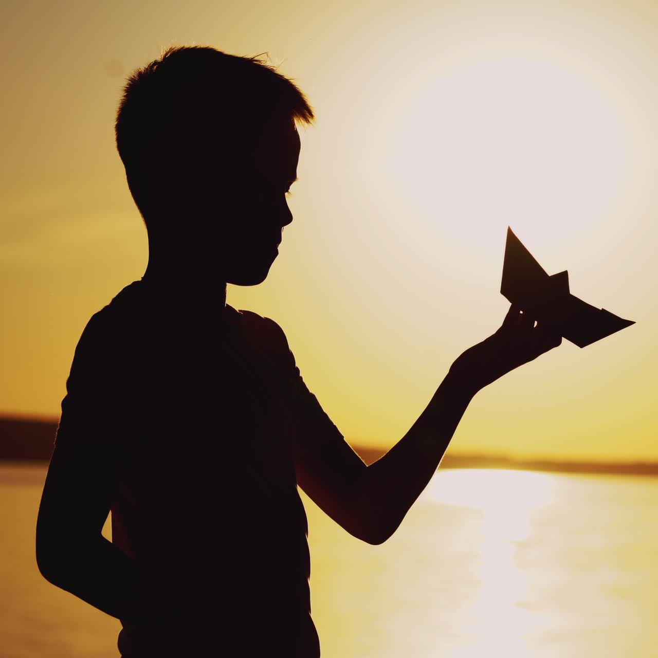 Silhouette of a child by the river. Paper boat is in the hands of the child. Beautiful summer sunset. Origami