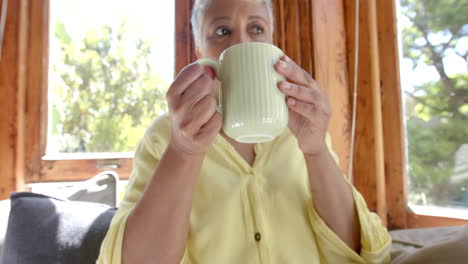 Happy-senior-biracial-woman-sitting-on-couch-and-drinking-tea-at-home,-slow-motion