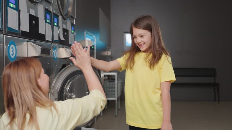 Back view of shopkeeper seated by washer loading clothes while young girl swings fabric playfully before keeping items, pair share high five in laundromat, teamwork mood, family moment