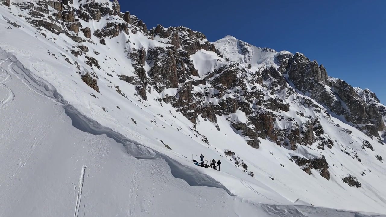 Un drone sale dal fondo di un pendio innevato, rivelando un gruppo di sciatori riuniti su un'alta cresta montuosa con l'imponente vetta del Monte Tetnuldi che si erge sullo sfondo in Svaneti, Georgia